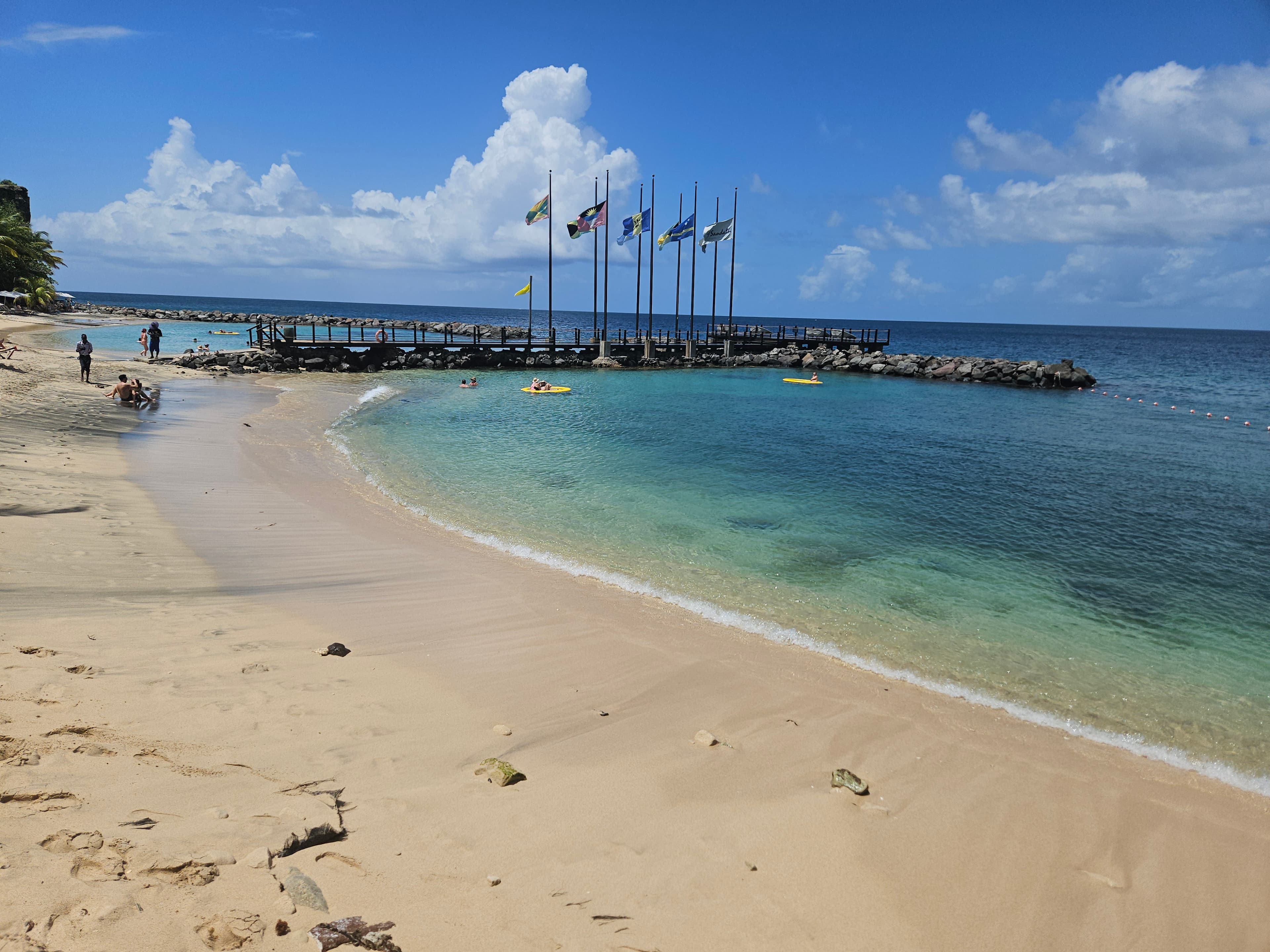 View of a calm sandy beach with a rocky pier with flags flying above on a sunny day