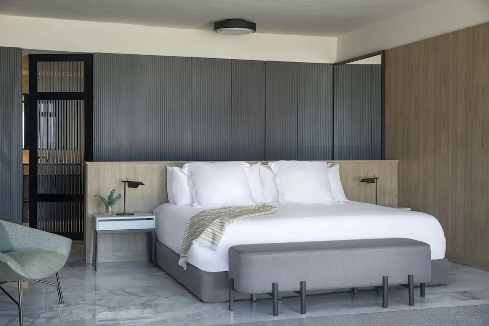 A modern hotel room with grey walls and floors, white bed linens, and light brown wood.