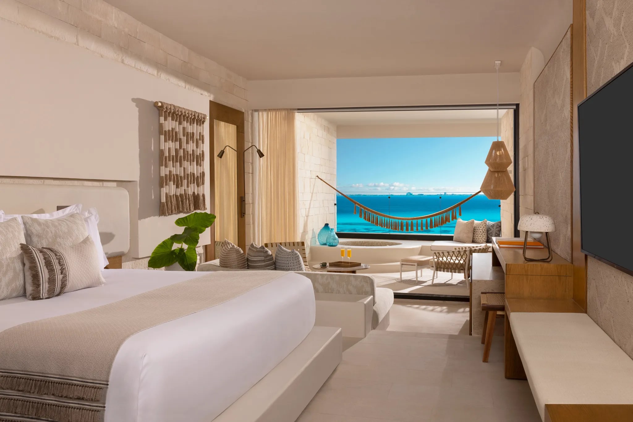A hotel room with a bed (covered in white linens and tan decor), tan walls, and a wall mounted television leading out to a balcony with a hammock and bright turquoise ocean in the distance
