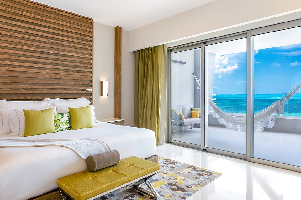 A hotel room with brown wood, white linens, and bright yellow & green decor, leading out to a balcony with white hammock and bright turquoise ocean in the distance