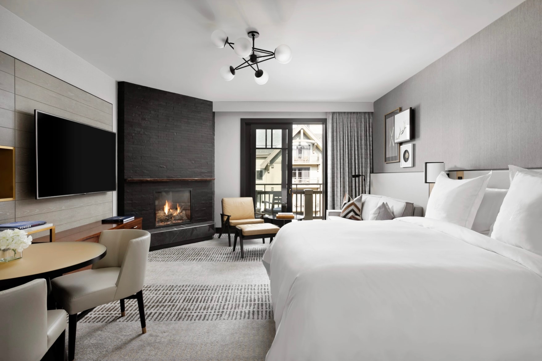 A hotel room with white bed linens & walls, light grey rugs and a black fireplace with a lit fire