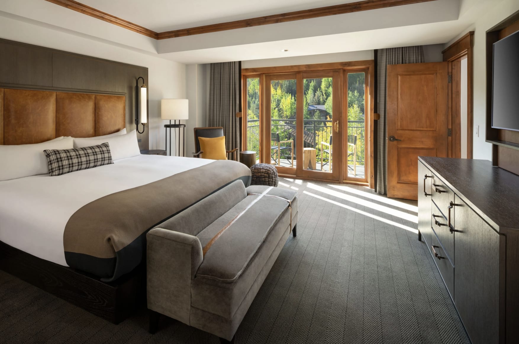 A modern rustic hotel room with brown wood, white walls, grey carpets and white bed linens with a forest outside the windows in the background