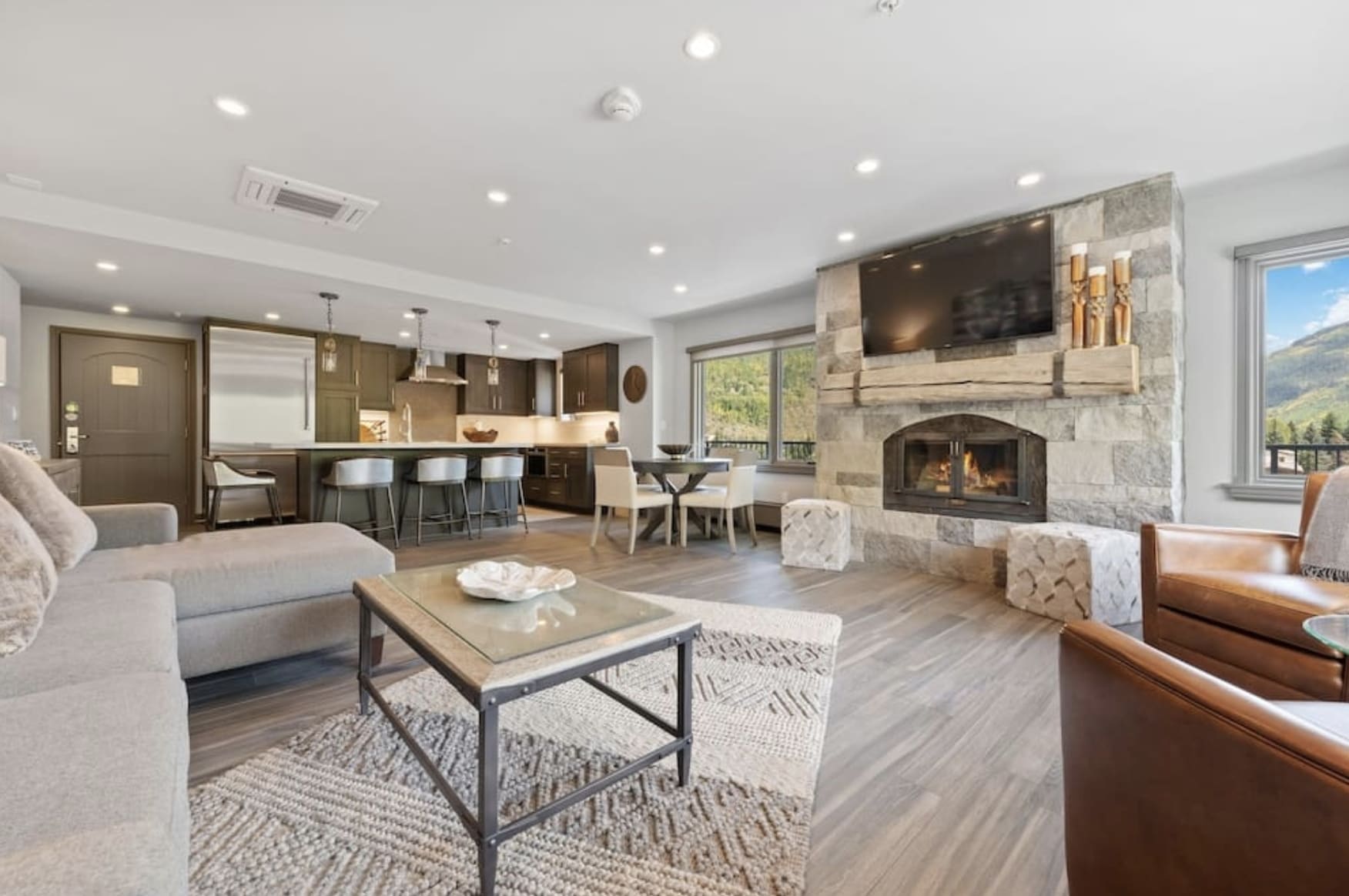 A living room with light colored decor, stone fireplace, and modern kitchen in the background