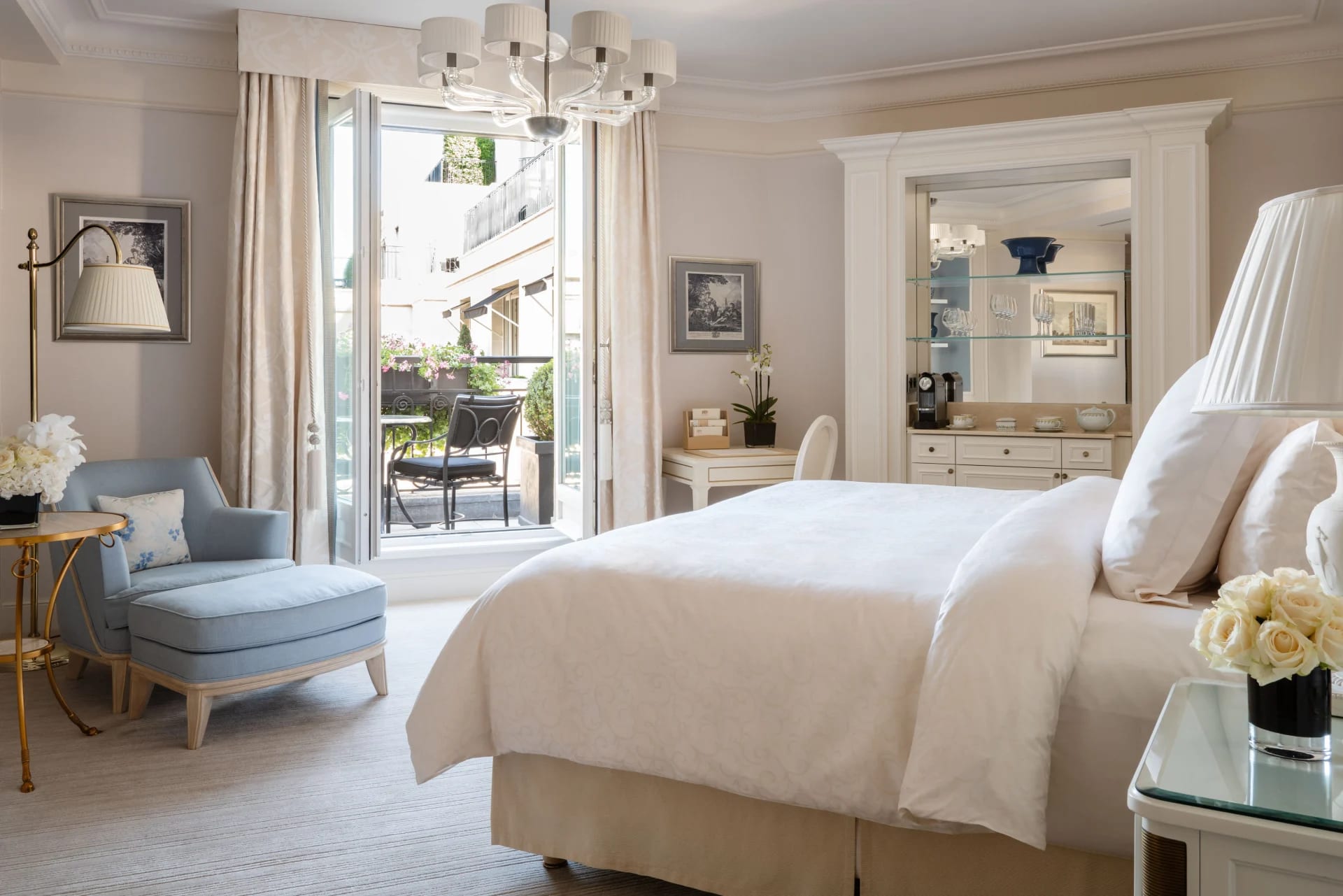 A side view of a hotel room with white linen covered bed on the right, a light blue sitting chair on the left, and open French doors onto a balcony in the back
