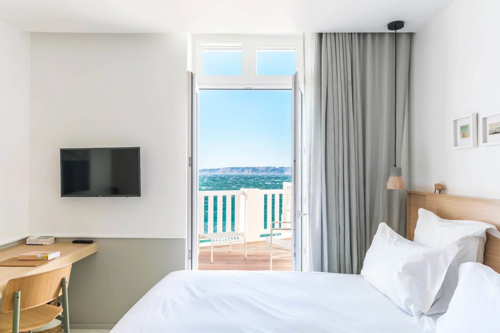 A white bedroom with light wood furniture, white linens and light grey curtains leading out to a balcony overlooking bright turquoise ocean in day