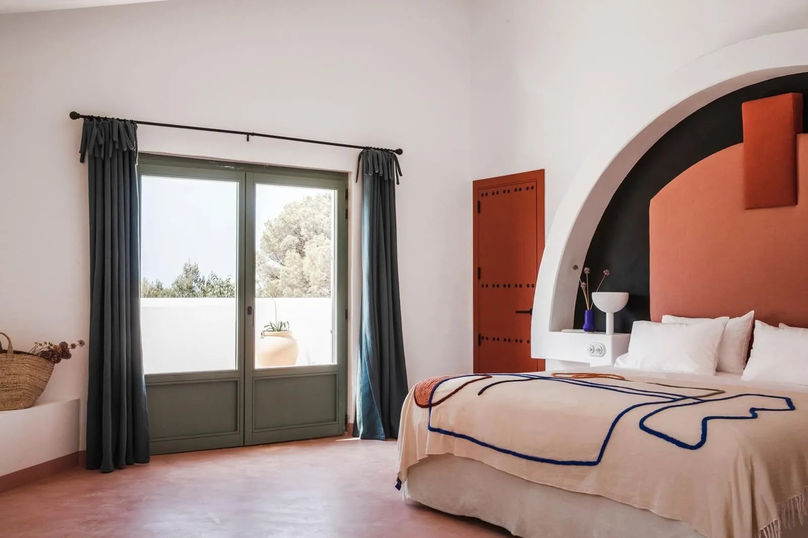 A modern hotel room with brown wood floors & door, a bed with modern pattern bedspread, orange curved headboard, and green curtains framing glass doors leading outside