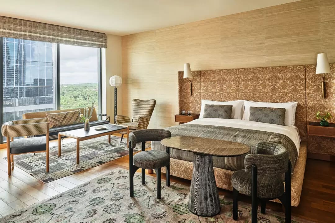 View of modern hotel room with tan and grey tones, table and chairs at the foot of a bed with a side seating area next to a window