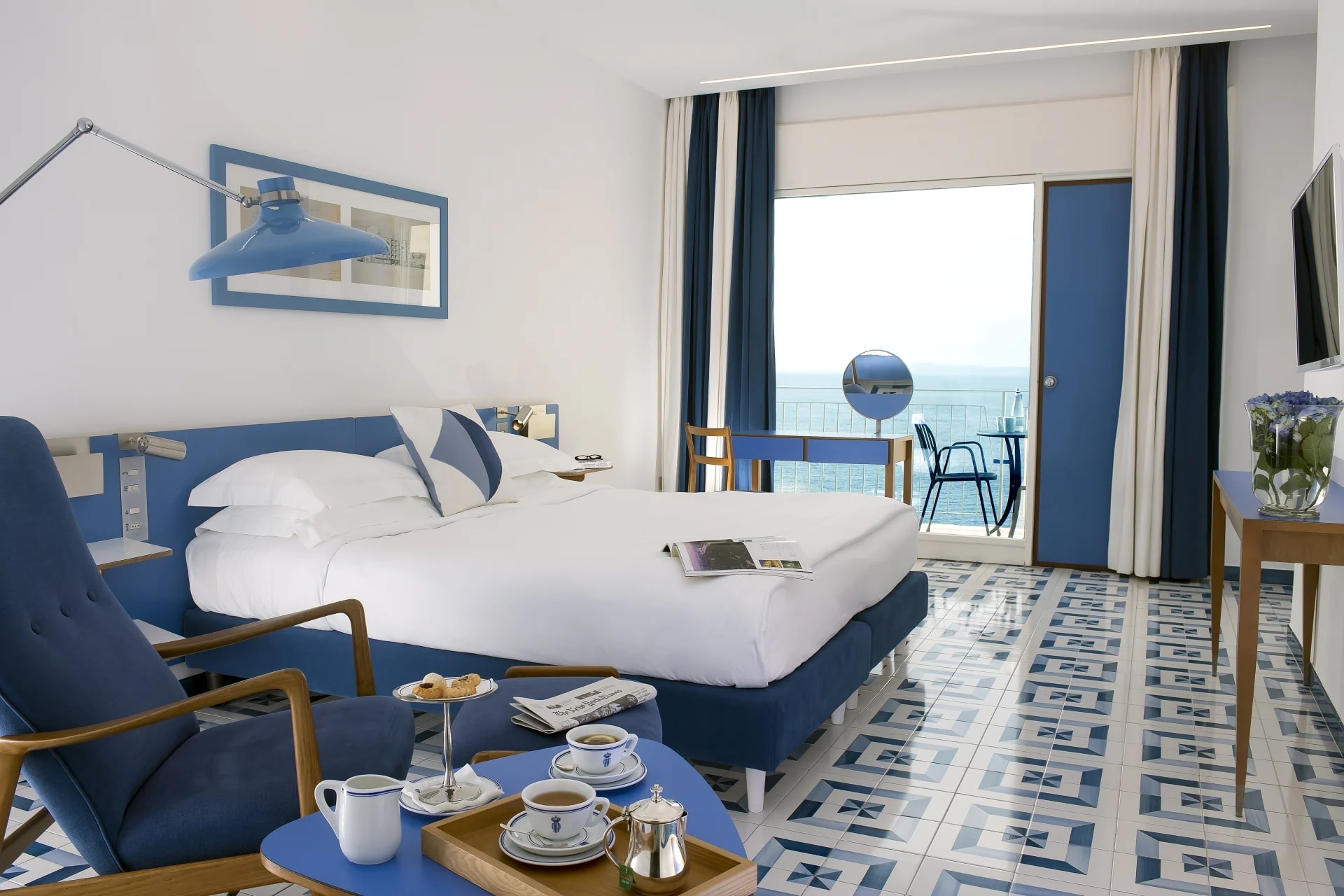 A hotel room with blue armchair, white sheet covered bed, blue headboard, white walls, white and blue tile floor, and open balcony with ocean view