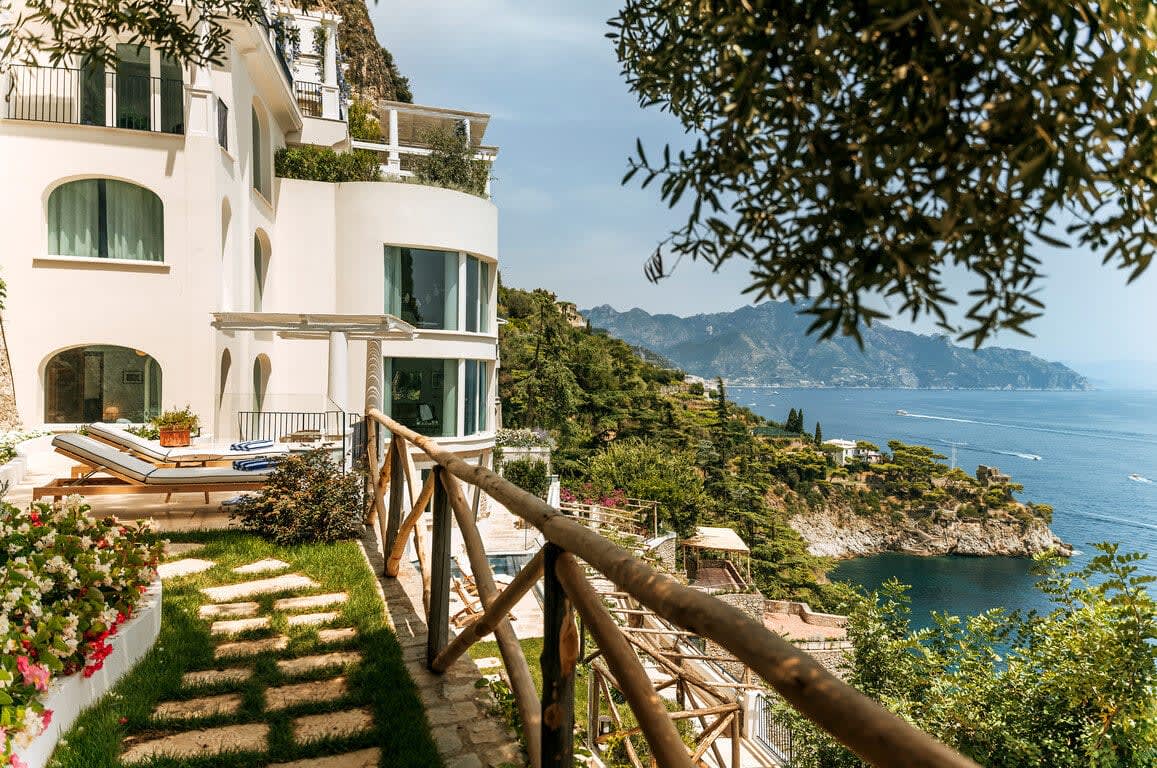 Green grass covered terrace with lounge chairs and a white building and wood railing with hills and ocean beyond during day