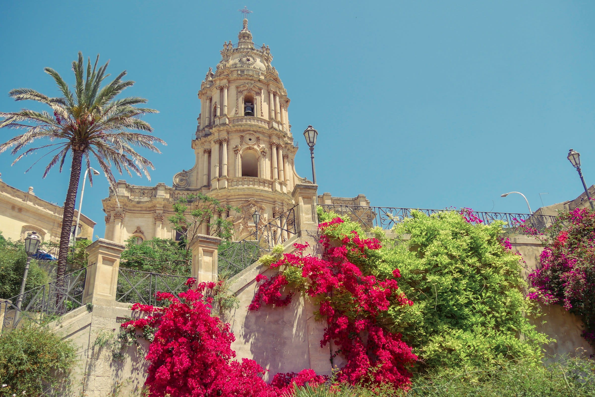 View looking up at tan cathedral tower with bright flowers, green bushes and palm tree