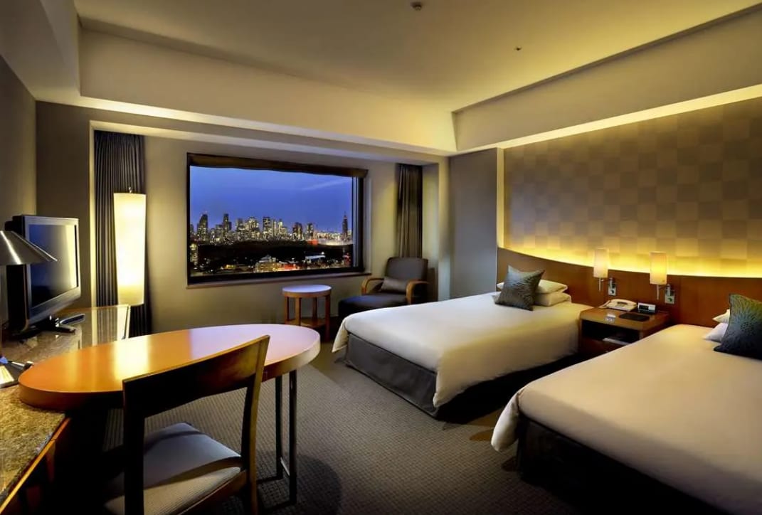 A hotel room with two beds, gray carpet and a window showing Tokyo cityscape