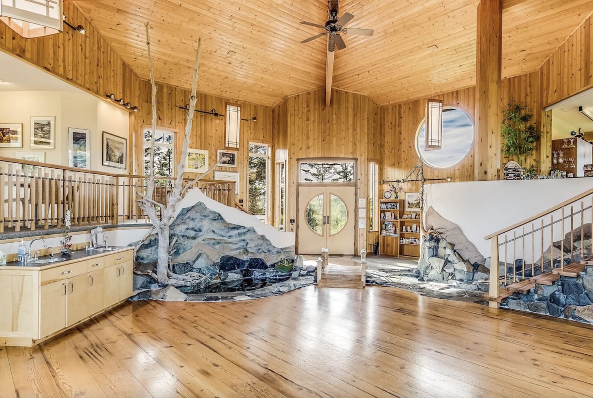A cabin interior with wooden floors and ceilings, as well as stone stone stairwells, ceiling fan and trees decorating the interior