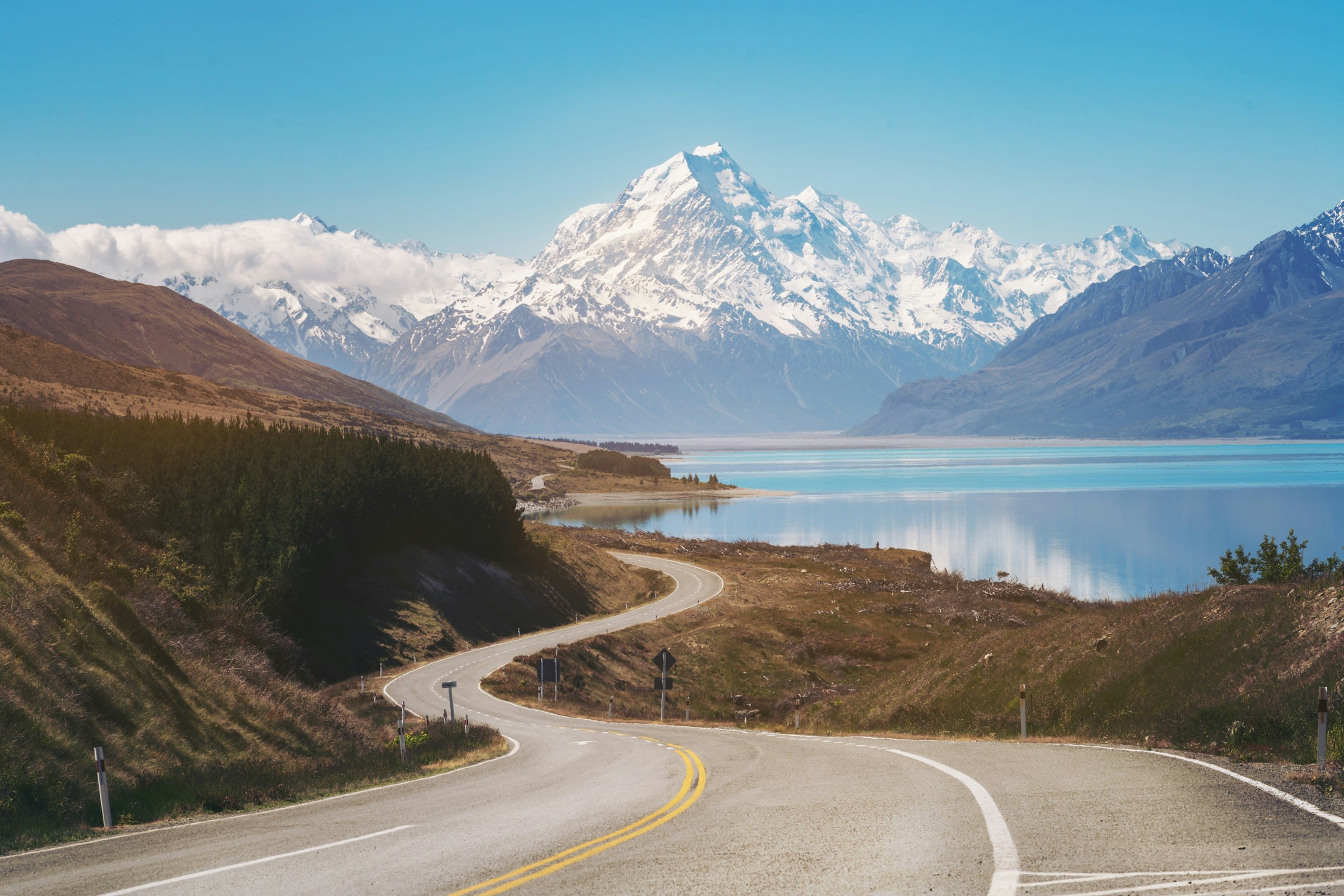 A winding road leading to a lake and a view of snow covered mountains