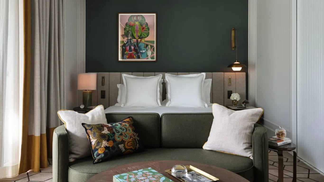 A green loveseat in front of a white linen covered bed and green walls