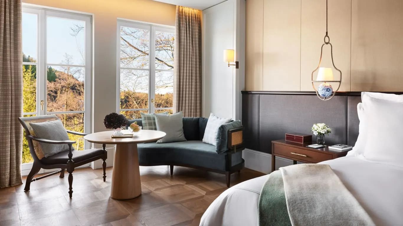 A hotel room with a bed off to the right side, light tan walls, a green loveseat and chair with side table in front of floor-to-ceiling windows