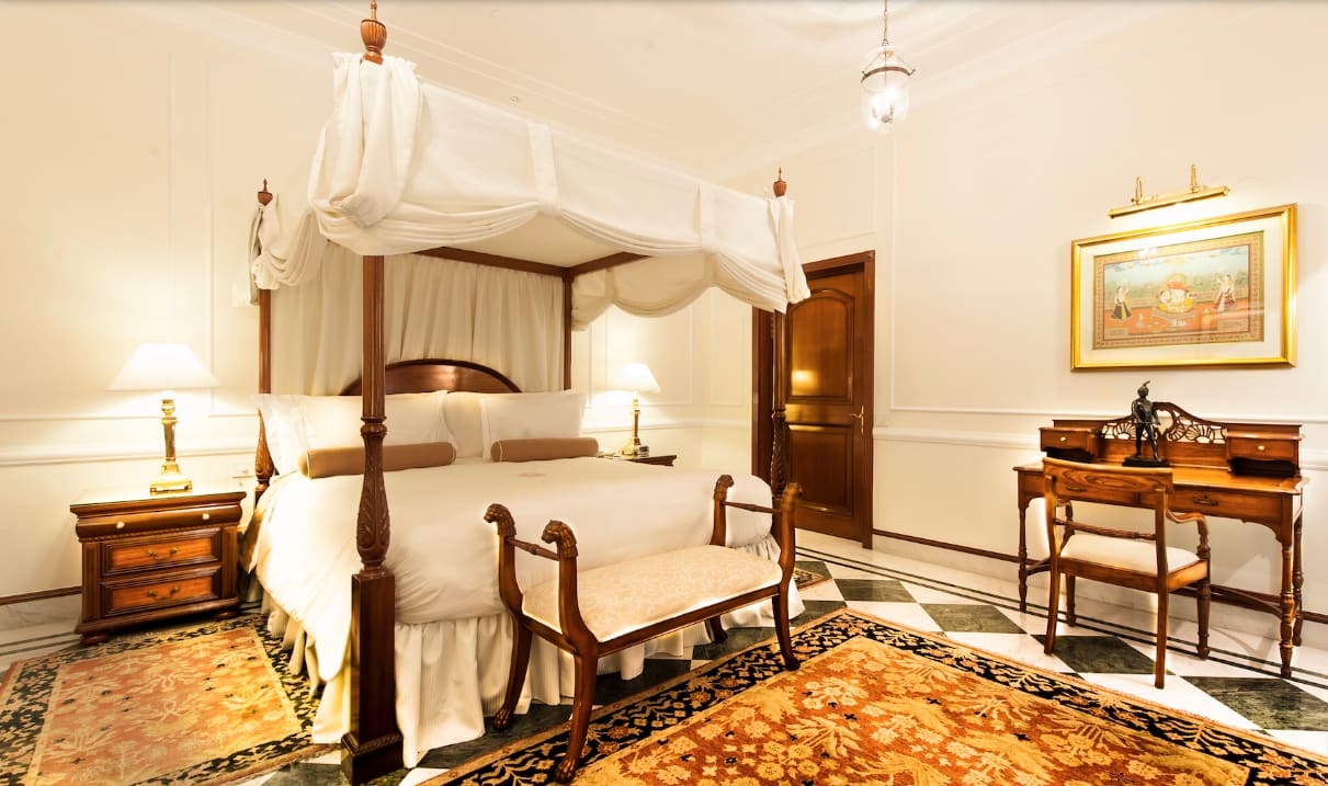 A white hotel room with orange, tan and black decorative carpet, dark wood four poster bed and dark wood desk