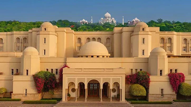 A light tan stone building entrance with vibrant green forest behind it and white stone landmark on the horizon