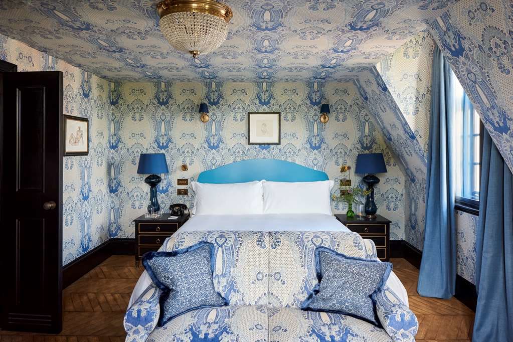 A traditional English hotel room with white and blue patterened wallpaper, blue curtains, and white bed linens