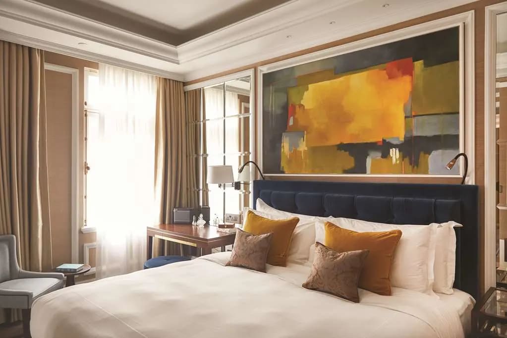 A contemporary hotel room with white bed linens, brown pillows, tan curtains, and multicolored painting above the bed