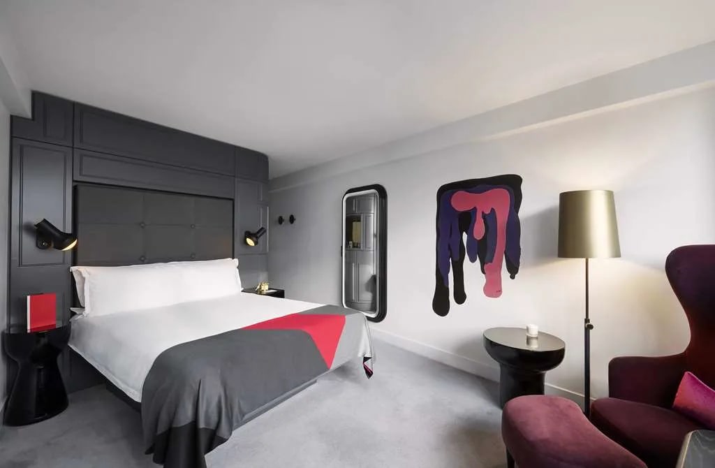 A modern hotel room with dark grey accent wall behind the bed, red accents and red side chair