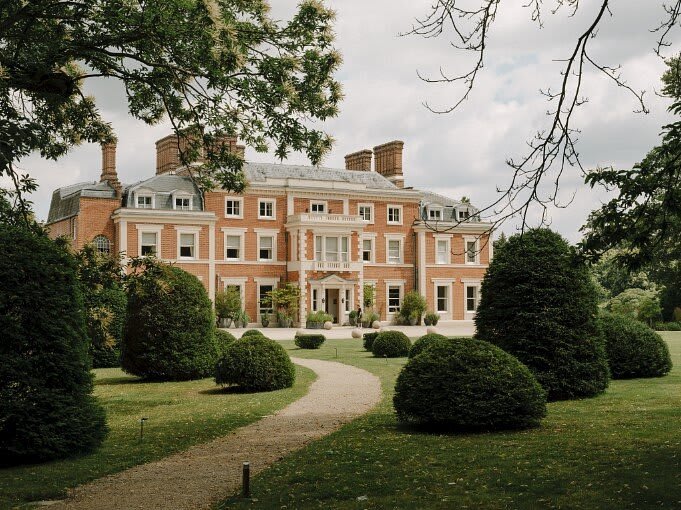 A landscape view of manicured lawns and trees with a dirt path leading to a grand brick country house