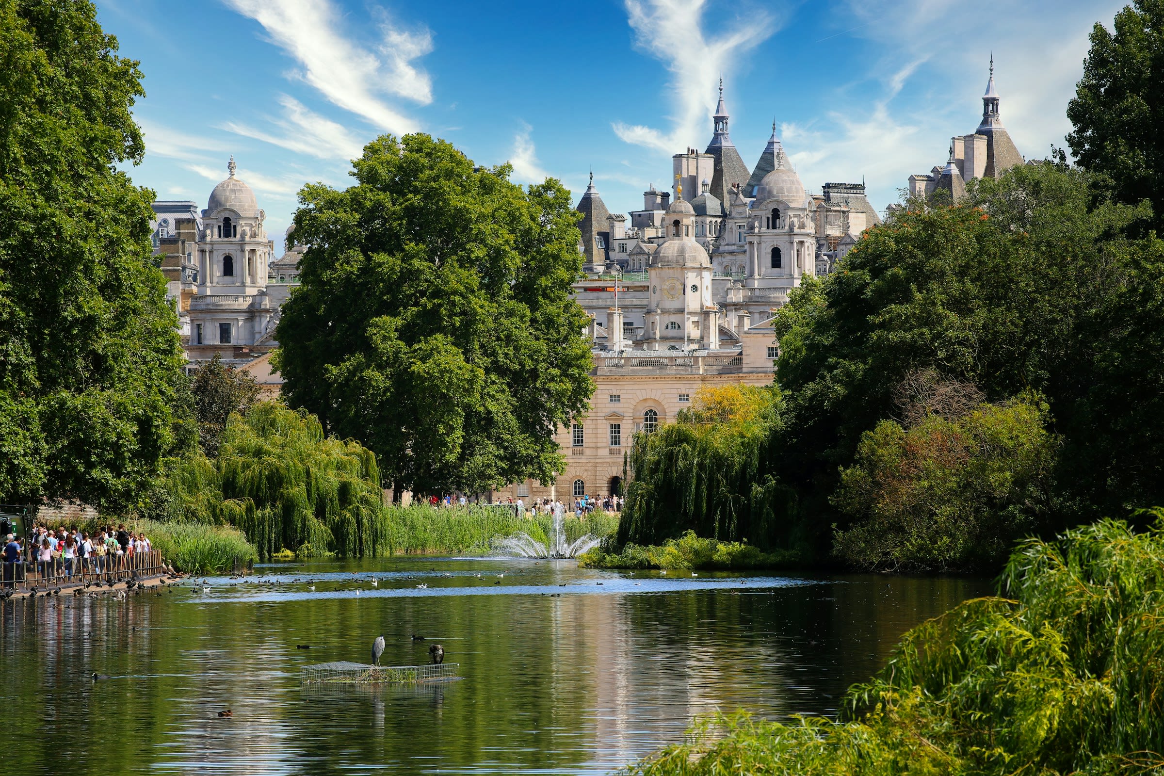 A lake in a green park with English traditional buildings behind during daytime