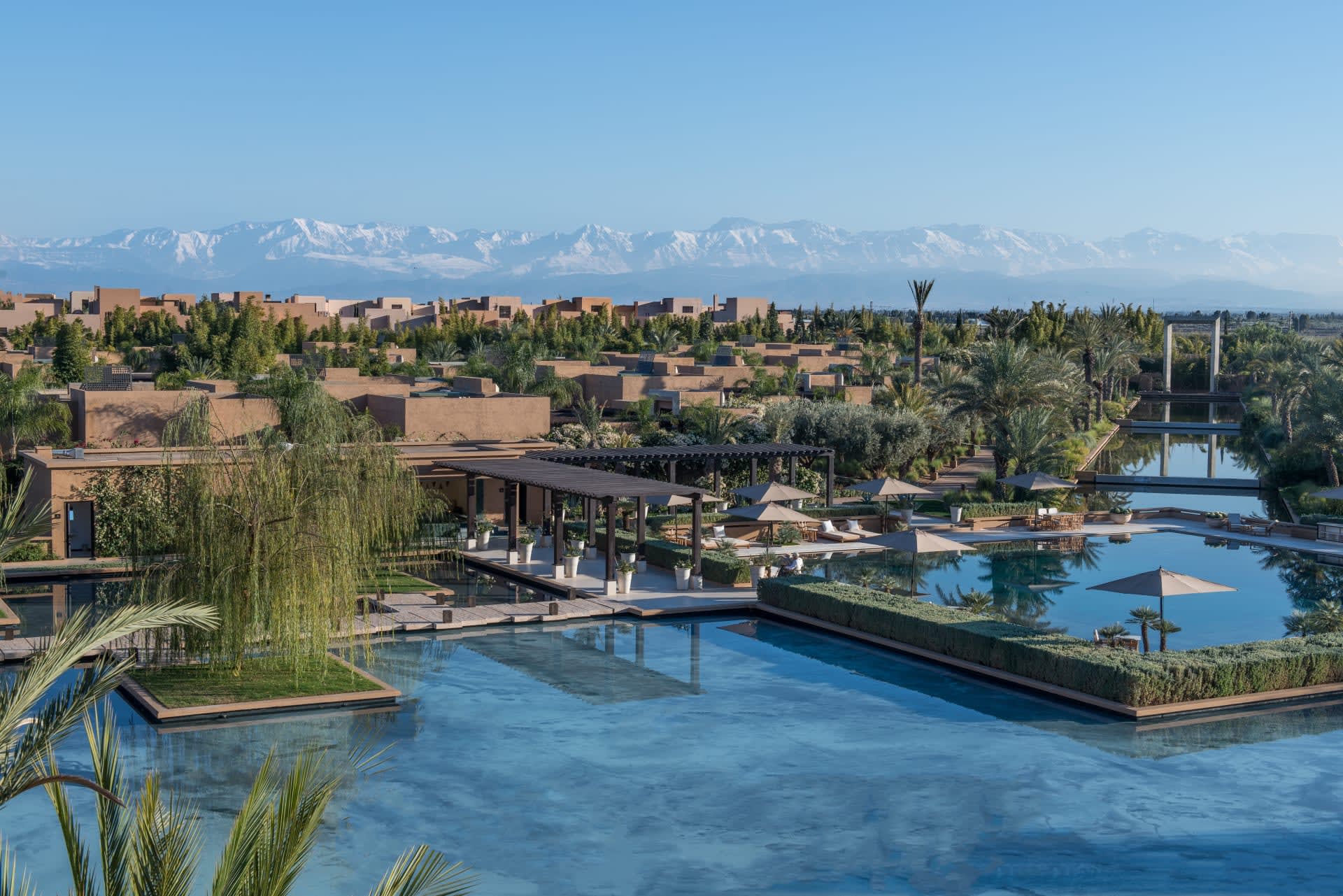 Aerial view of expansive pools amid landscape greenery and tan buildings with snow capped mountains in the distance