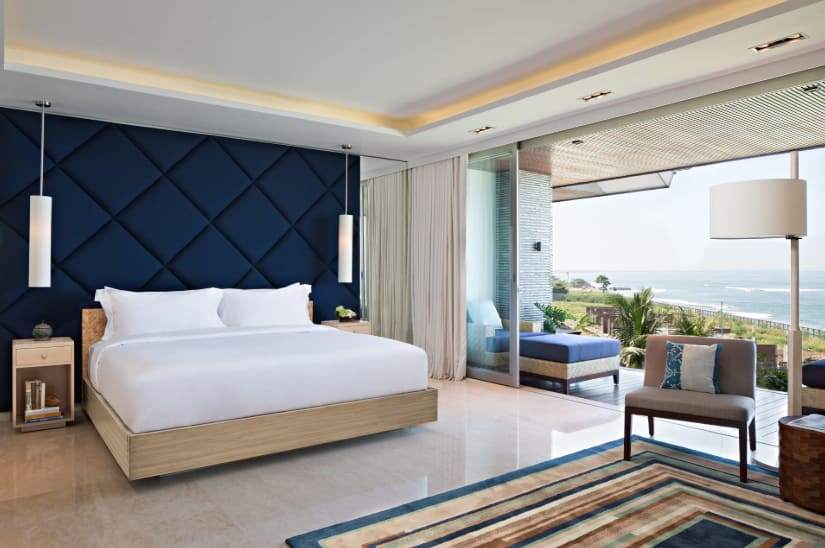 Modern room with blue tufted wall behind a white linen bed, natural wood furniture leading out to a terrace with sea view