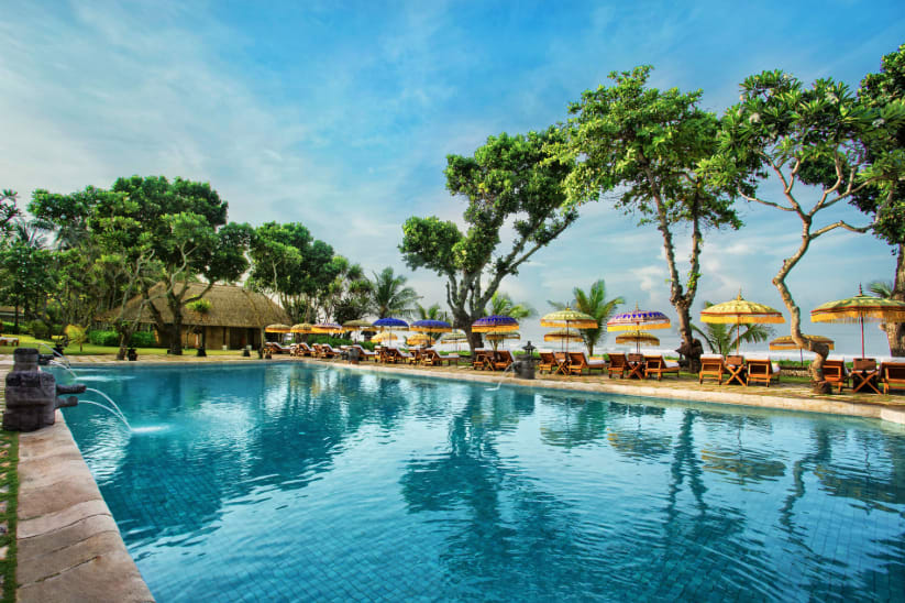 A turquoise swimming pool lined with wooden lounge chairs and ornate umbrellas along the beach surrounded by tropical trees