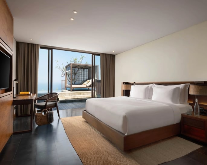 Brown and white room with white bed linens, low sitting wood furniture leading out to a lounger on the terrace with tree
