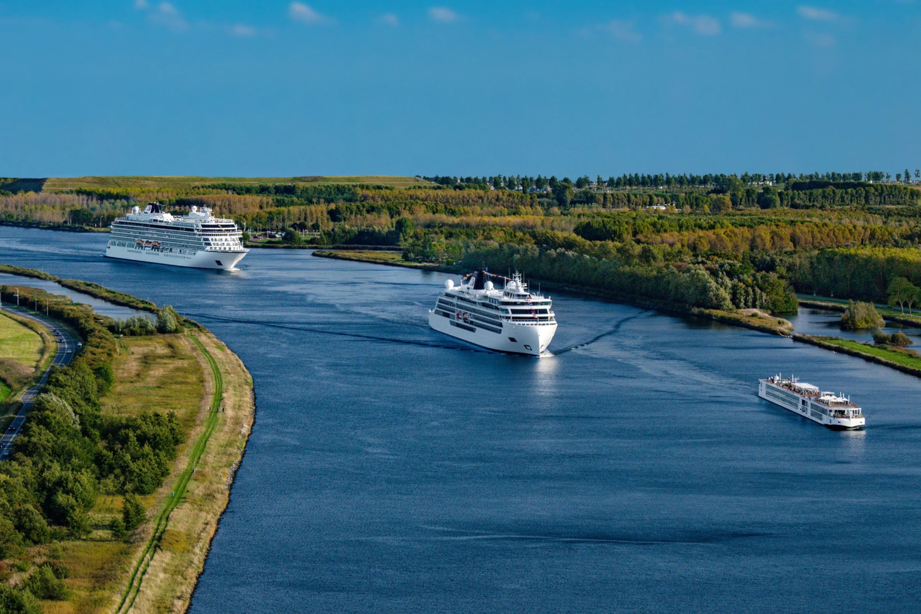 Three ships in decreasing size sail along a calm river with greenery on either side