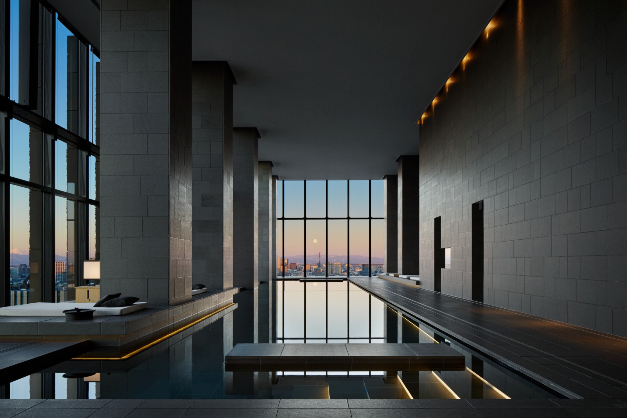 an indoor swimming pool in a sleek room with tall ceilings and large windows overlooking a city