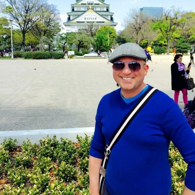 Travel Advisor Travis Tanner wears a blue sweater, cross body bag, sunglasses, and a beret hat in front of a Japanese temple. 