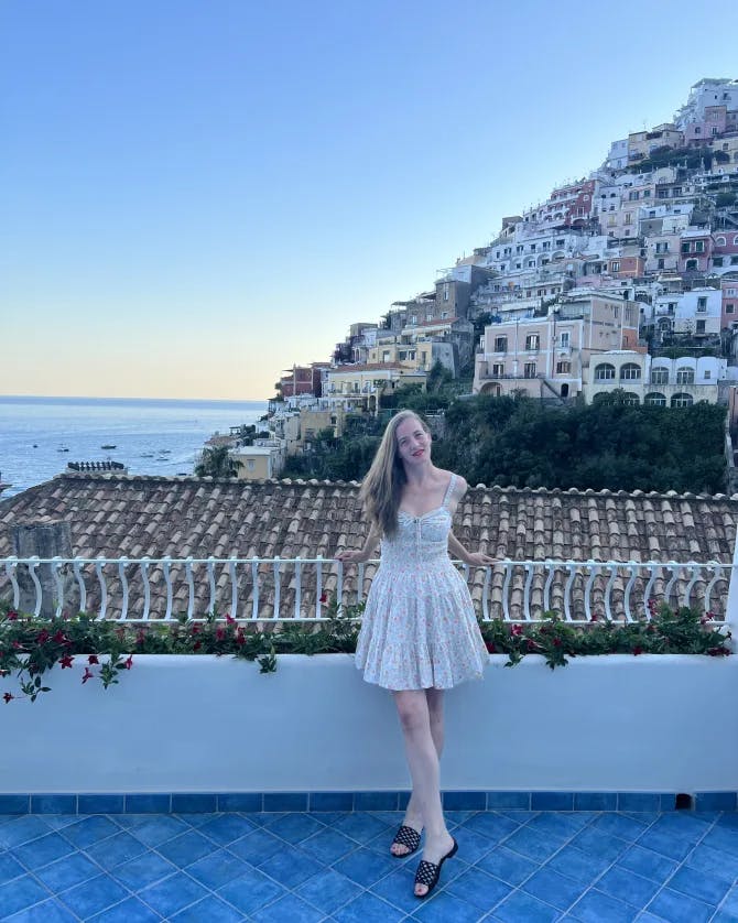 A picture of Sonia wearing a white floral dress and standing on a balcony in front of an Italian coast with the sea in the background 