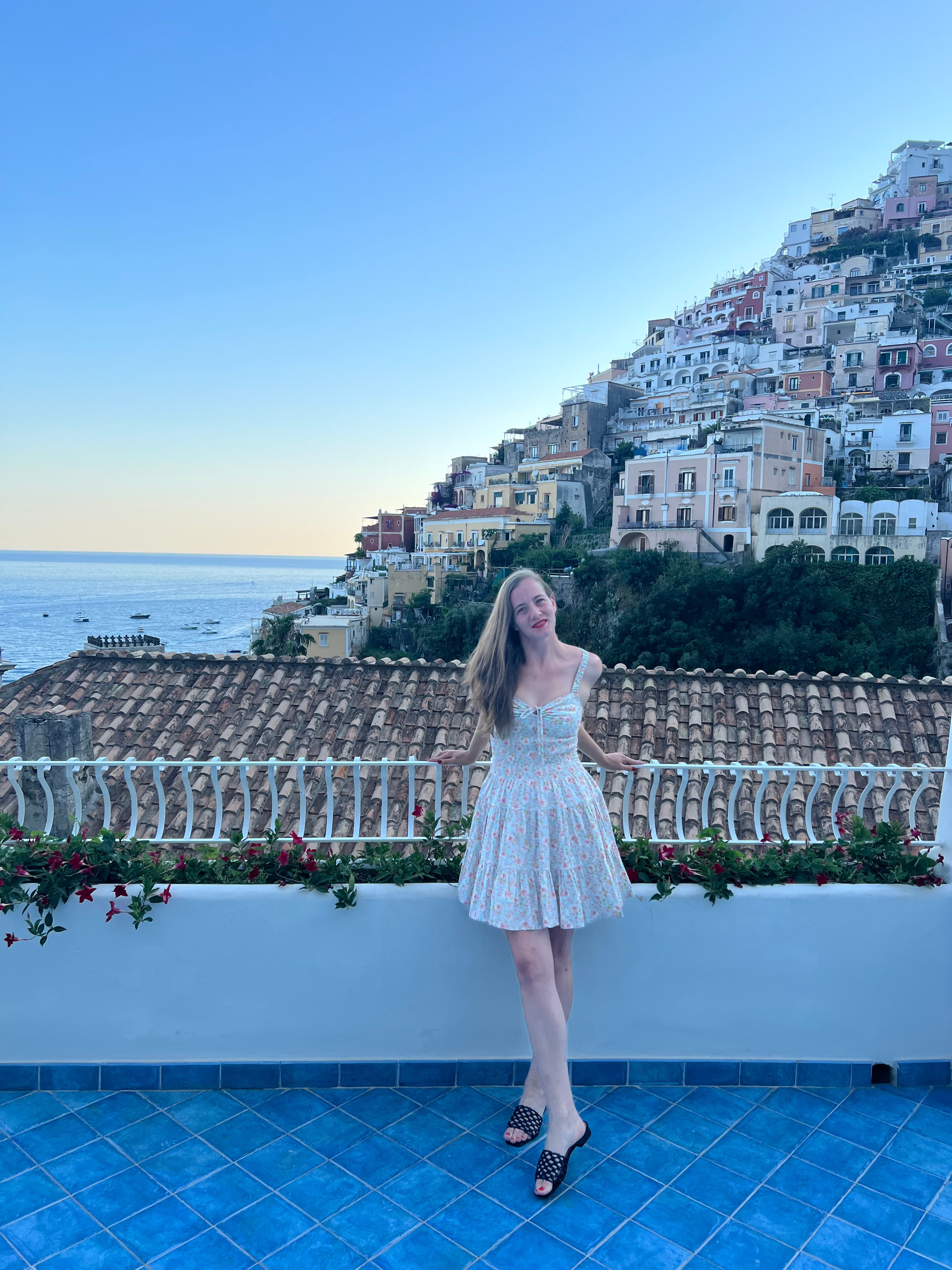 A picture of Sonia wearing a white floral dress and standing on a balcony in front of an Italian coast with the sea in the background