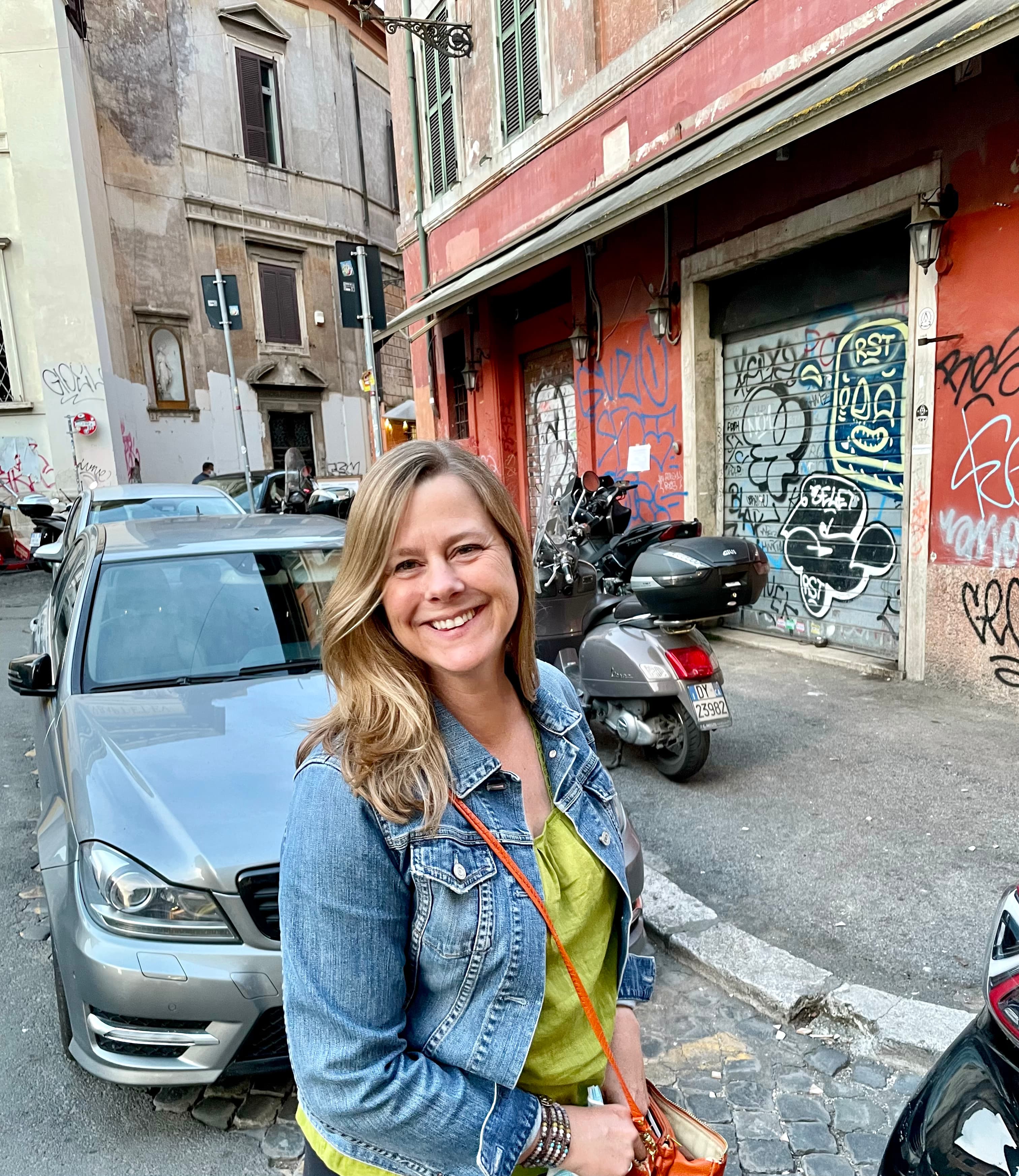 Jennifer wearing a green top and denim jacket standing outside on a city street in front of a car, motorcycle and red building covered in graffiti