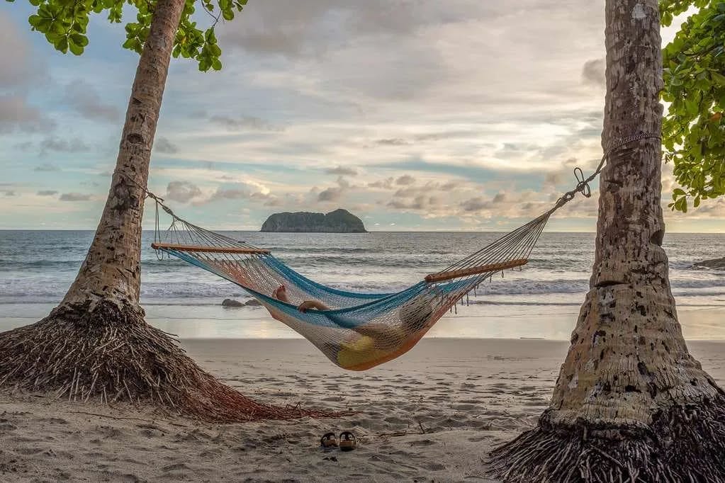A man in a swimsuit and tank top lounges in a colorful hammock strung between two trees at a Manuel Antonio beach