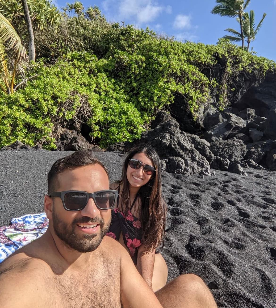 Khushbu and friend sitting on a tropical, black-sand beach, smiling and taking a selfie.