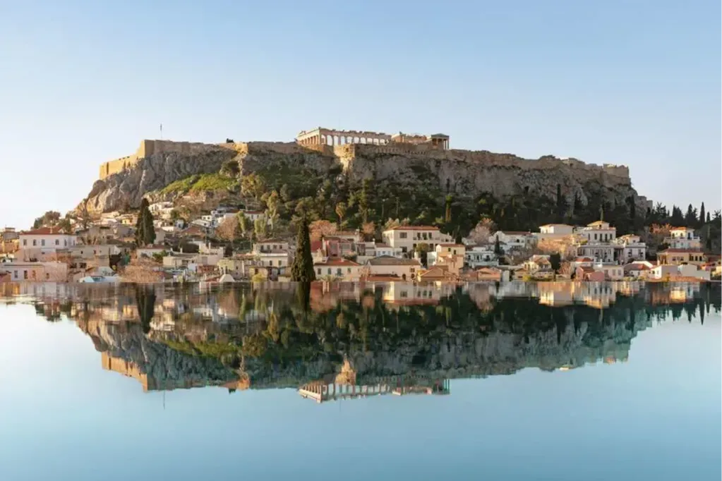 The Acropolis stands above historic structures lining the shores of Athens, which are mirrored in the calm waters