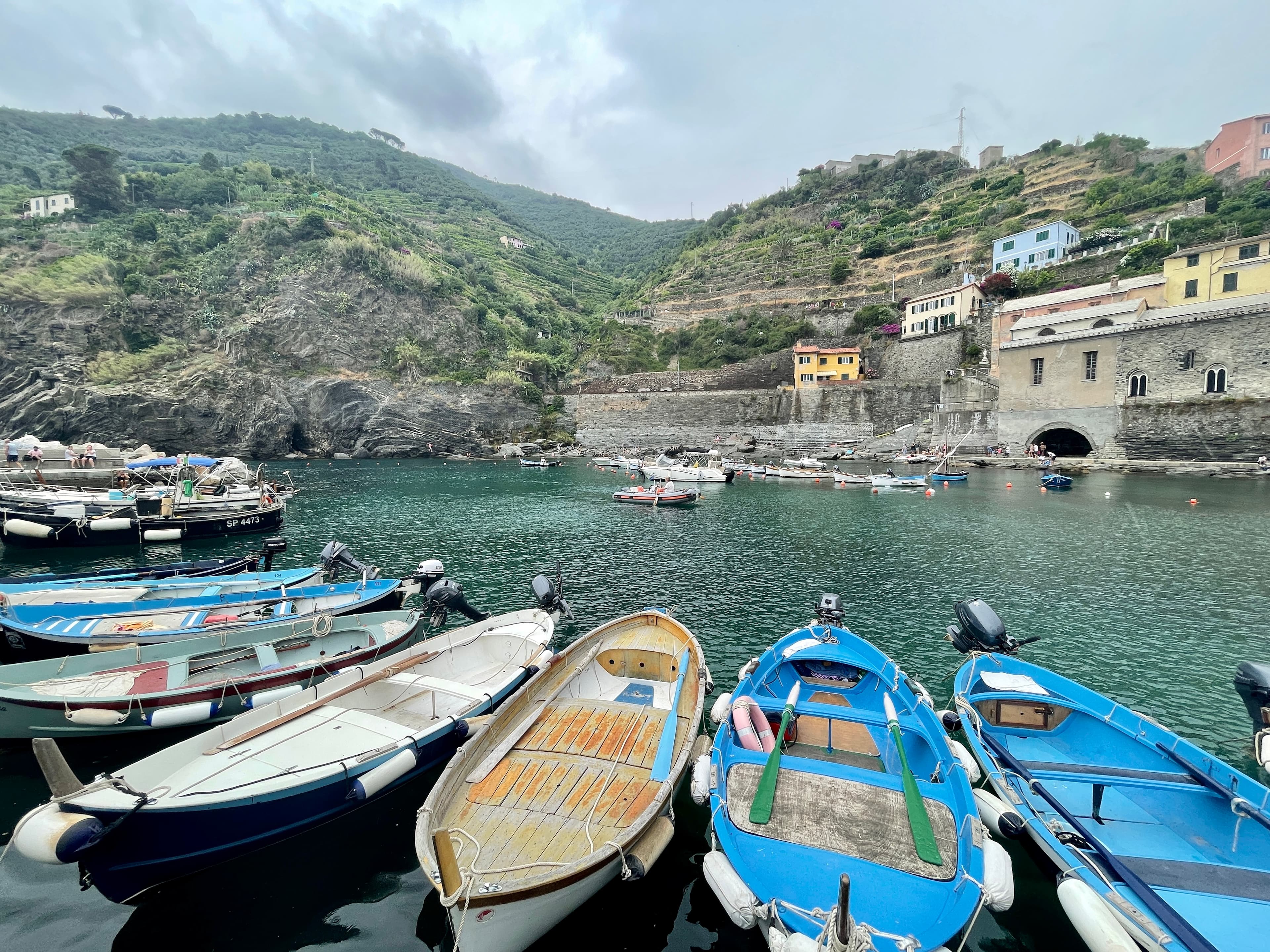 Six boats that are blue, white and tan docked in front of turquoise blue water and buildings set into a rocky mountain scattered with trees