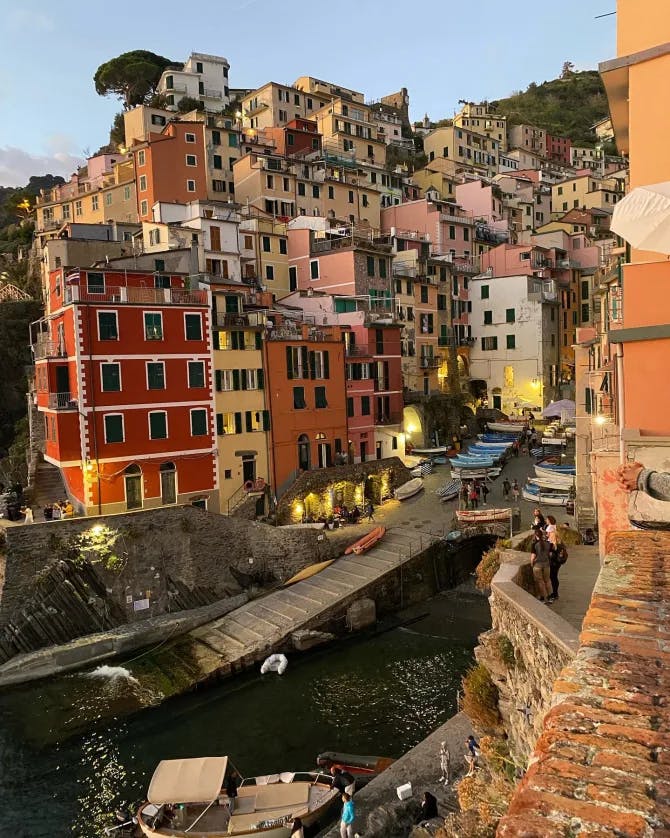 A beautiful view of the colorful buildings in Italy nestled around a harbor with stone pathways and tourists walking around. 