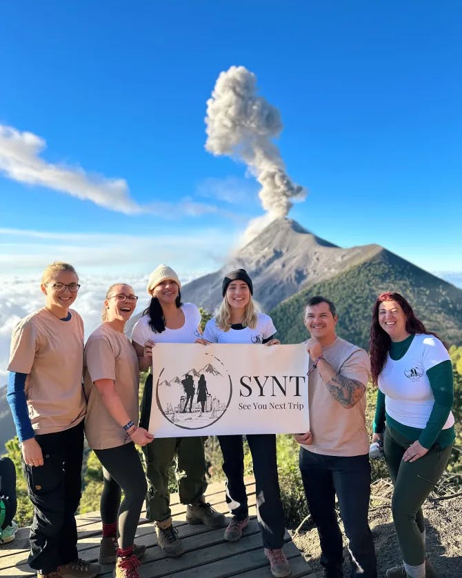 A group photo of six people holding up one sign and standing in front of a volcano