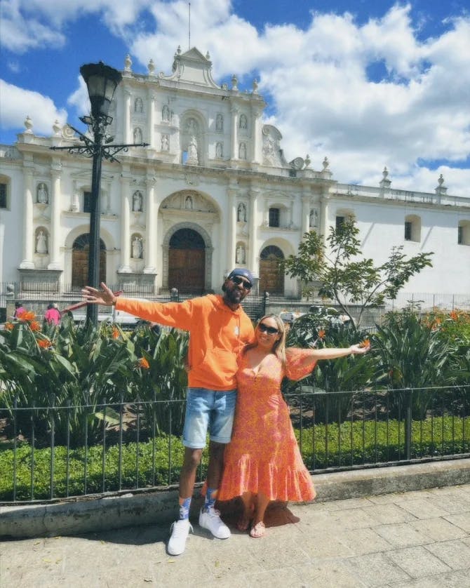 Layne and his partner wearing orange clothing and posing with their arms stretched wide while visiting Antigua, Guatemala Cathedral