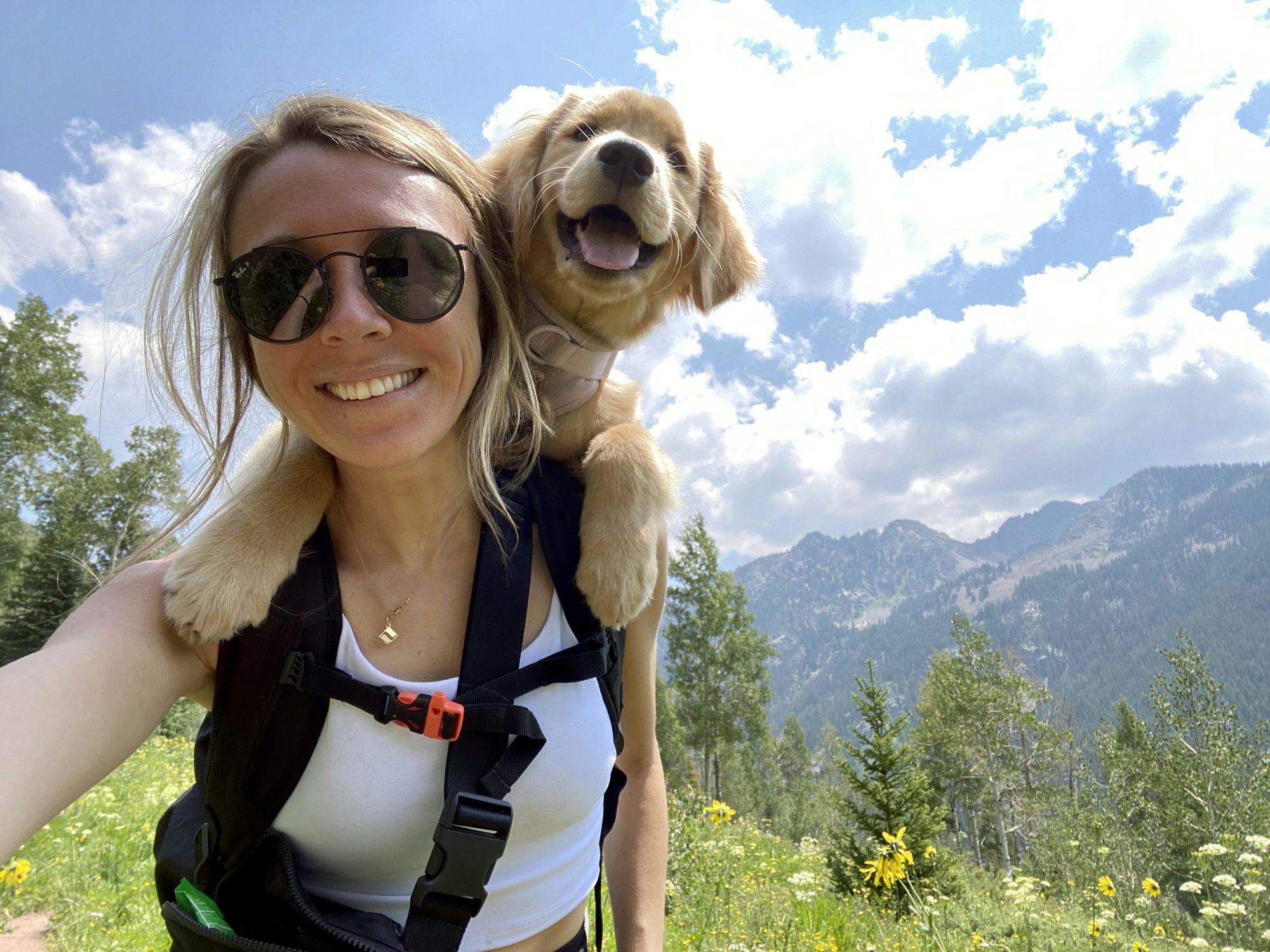Travel Advisor Charlie Gaffney smiles at the top of a wild flower mountain with a black backpack and a dog smiling over her shoulder