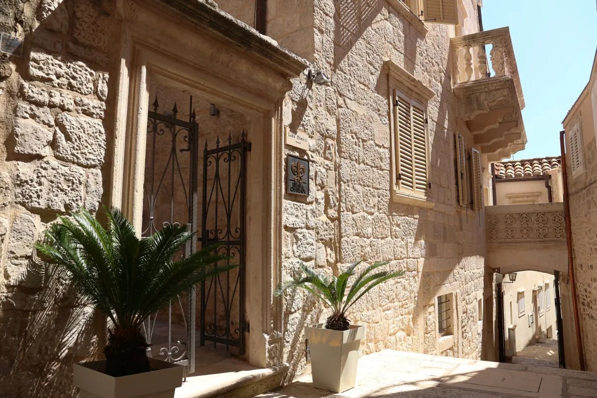 A beautiful stone building with a metal gate door, palm trees in pots and an archway beneath a balcony to the right.