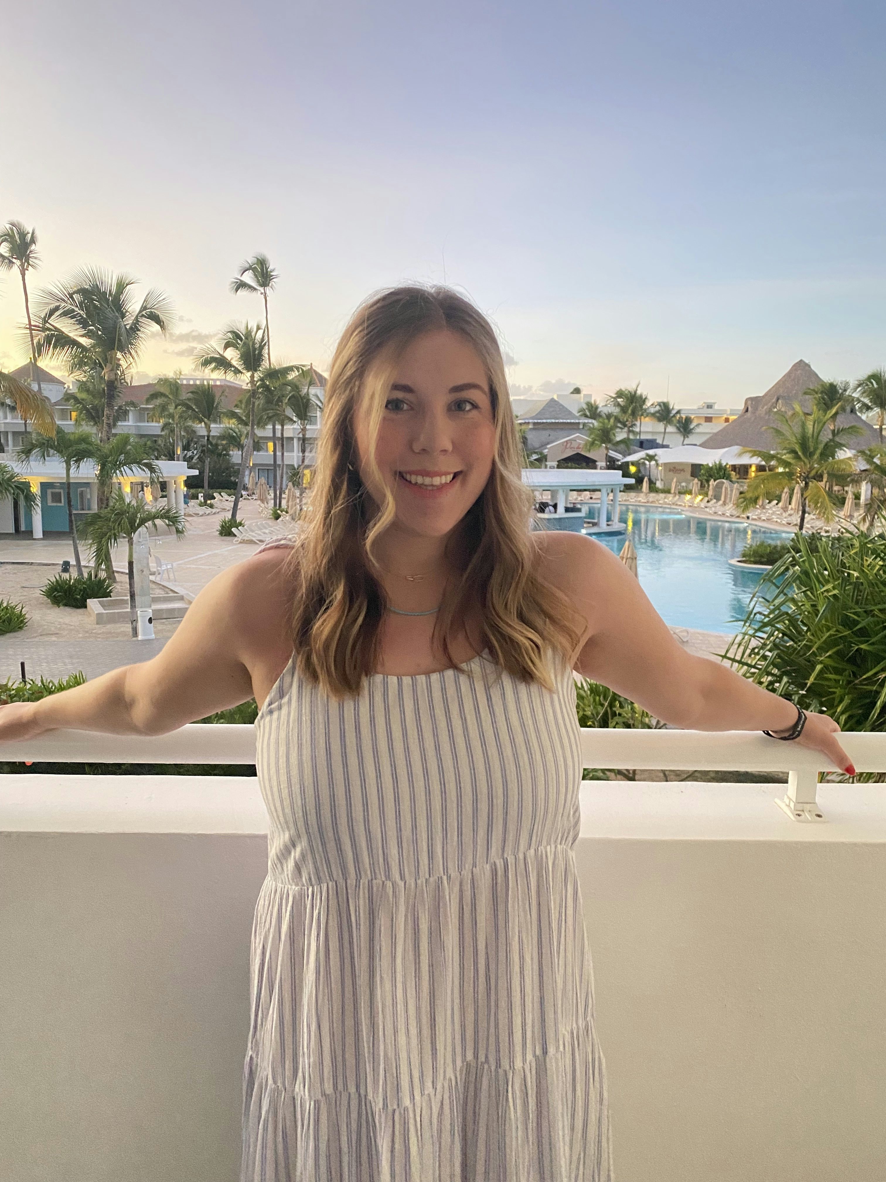 Travel Advisor Mallory Ashmore smiles in a white dress on a scenic, poolside balcony.