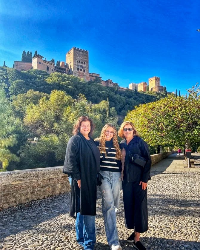 Luanna and two friends posing on a cobblestone road with trees and a castle on a hill in the background