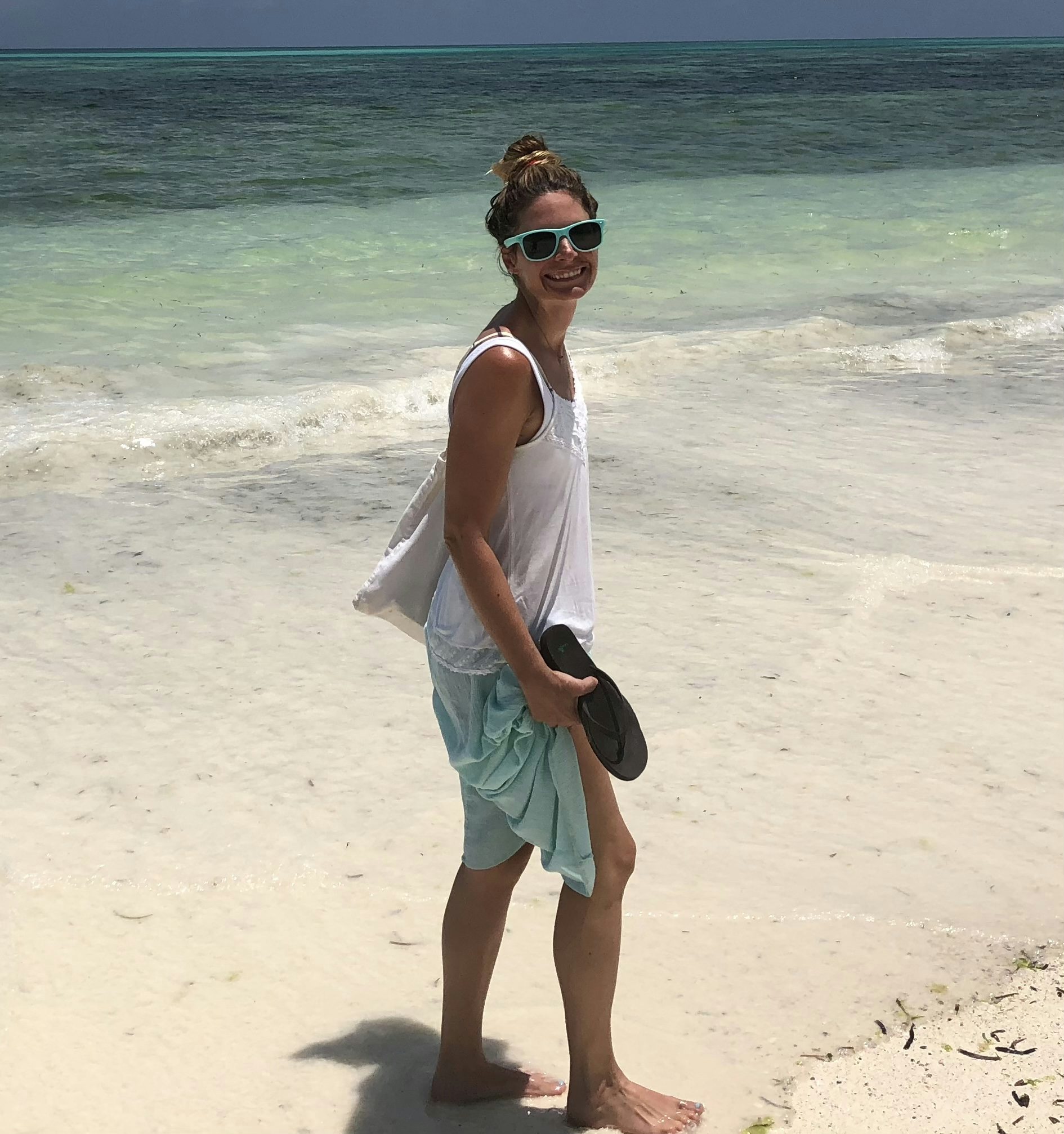 Travel advisor Keeley Landes smiles on a beach wearing a white tank top and turquoise sunglasses