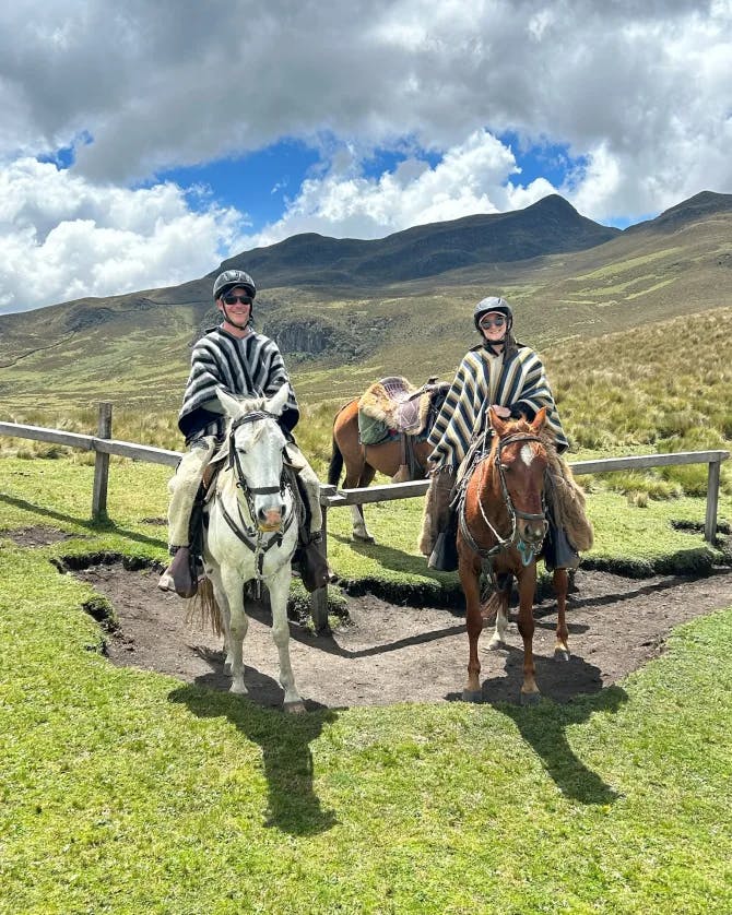 Stephen and his partner each riding a horse while wearing a black and white striped poncho outside on a green meadow with a mountain range in the background