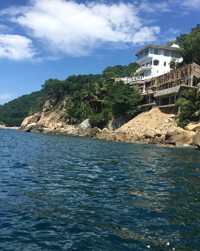 Blue sea water next to trees and a large white house tucked into a rocky coastline 
